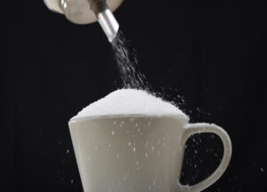 man hand with sugar bowl pouring a crazy lot of it spilling out everywhere in full coffee cup in insane sugar addiction and unhealthy nutrition concept isolated on black background ** Note: Shallow depth of field