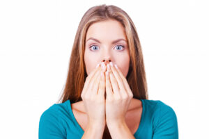 Surprised young woman covering mouth with hands and staring at camera while standing isolated on white